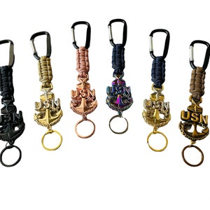 May include: Five keychains with a US Navy anchor charm. The keychains are made with paracord and have a black, tan, rose gold, rainbow, and navy blue color scheme.