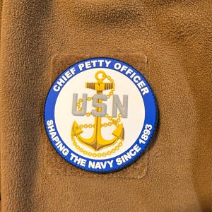 May include: A brown fleece jacket with a circular patch. The patch features a gold anchor, the letters "USN" in gray, and the text "CHIEF PETTY OFFICER SHAPING THE NAVY SINCE 1893" in blue and white.