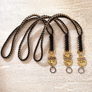May include: Three braided lanyards with black and tan cords. Each lanyard has a gold-colored anchor charm with the letters "USN" and a small ring. The lanyards are displayed on a light-colored, textured surface.