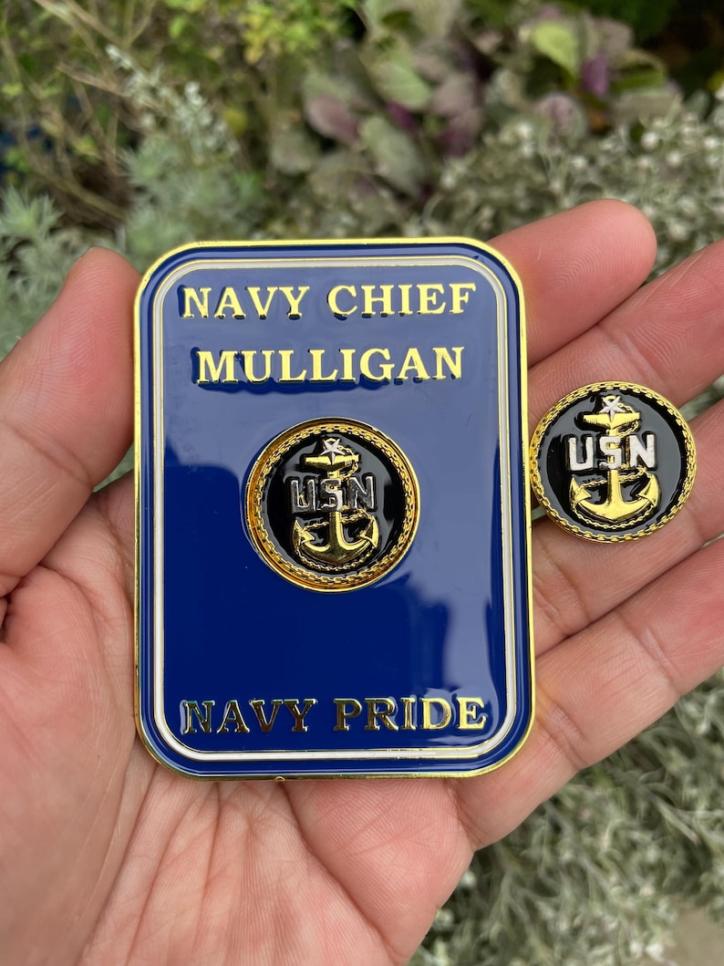 K&ouml;nnte beinhalten: Eine goldfarbene Metallnadel mit blauem Hintergrund und einem goldenen Anker mit den Buchstaben USN in Wei&szlig;. Die Nadel befindet sich neben einer gr&ouml;&szlig;eren goldfarbenen Metallplakette mit blauem Hintergrund und dem Text "NAVY CHIEF MULLIGAN" und "NAVY PRIDE".