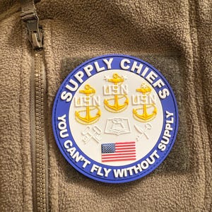 May include: A circular patch with the words "SUPPLY CHIEFS" in white lettering on a blue border. The patch features gold anchors, the USN insignia, and an American flag. The text "YOU CAN'T FLY WITHOUT SUPPLY" is also included.