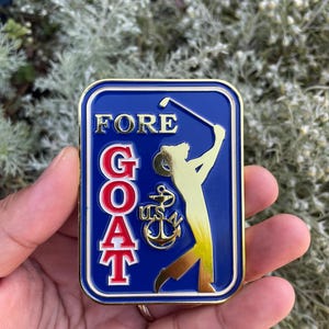 K&ouml;nnte beinhalten: Eine goldfarbene Metallstifte mit blauem Hintergrund. Die Stifte zeigt einen Golfer in einer goldenen Silhouette, ein rotes und wei&szlig;es Akronym "GOAT" und einen goldenen Anker mit der Aufschrift "USN" in goldenen Buchstaben. Die Stifte tr&auml;gt auch das Wort "FORE" in goldenen Buchstaben.