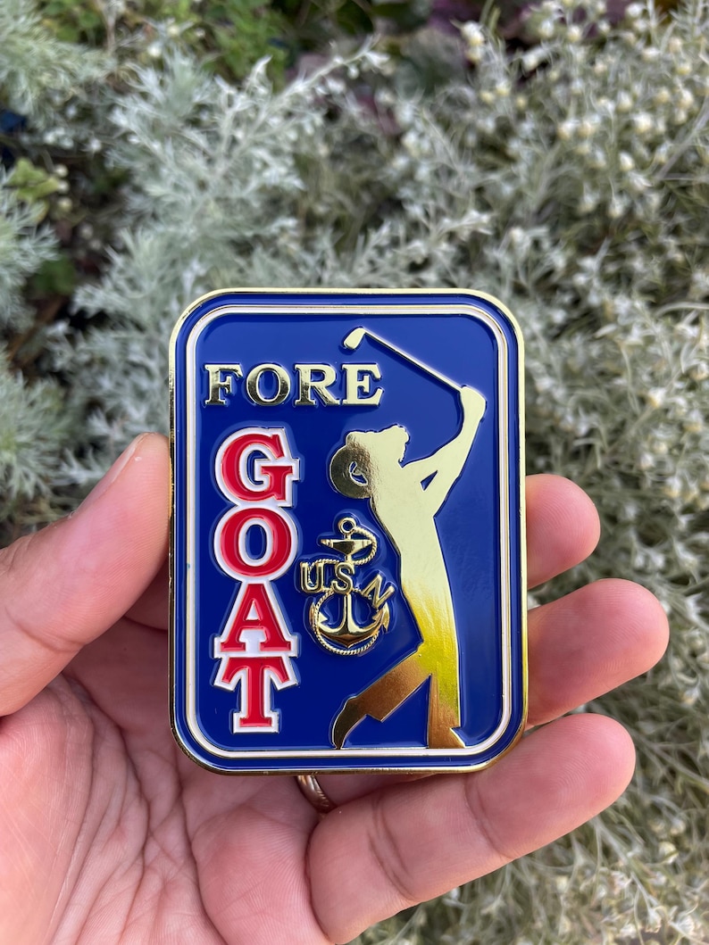 K&ouml;nnte beinhalten: Eine goldfarbene Metallstifte mit blauem Hintergrund. Die Stifte zeigt einen Golfer in einer goldenen Silhouette, ein rotes und wei&szlig;es Akronym "GOAT" und einen goldenen Anker mit der Aufschrift "USN" in goldenen Buchstaben. Die Stifte tr&auml;gt auch das Wort "FORE" in goldenen Buchstaben.