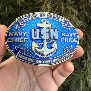 May include: An oval-shaped belt buckle featuring a blue diamond-plate background with a gold-tone anchor and chain. The text reads CLASS 132 FY 26, NAVY CHIEF, USN, NAVY PRIDE, and SHAPING THE NAVY SINCE 1893 in raised gold-tone lettering and borders.
