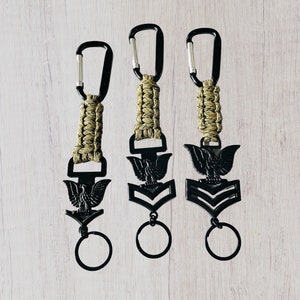 May include: Three black keychains with a braided cord and a metal eagle and chevron design. The keychains are on a light wood background.