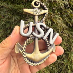 May include: A bronze and silver-toned anchor emblem with the letters "USN" and the text "CLASS 132 FY-26". The anchor is detailed with rope and is held against a backdrop of green foliage.