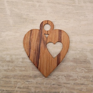 May include: Wooden heart-shaped pendant with a smaller heart cutout in the center. The pendant has a hole at the top for hanging. The wood grain is visible, with a mix of light and dark brown tones. The pendant is likely for jewelry or craft projects.