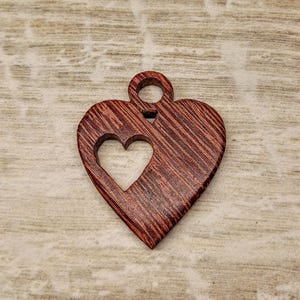 May include: A wooden heart pendant with a smaller heart cut out. The pendant is a reddish-brown color with a visible wood grain pattern. It has a small loop at the top for hanging.