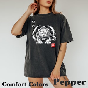 Puede incluir: Camiseta Comfort Colors gris oscuro con una ilustración blanca de un leopardo gruñendo y texto japonés. El diseño incluye un efecto de pincelada circular alrededor del leopardo y un cuadrado rojo con caracteres japoneses. La palabra "Pepper" está en la parte inferior.