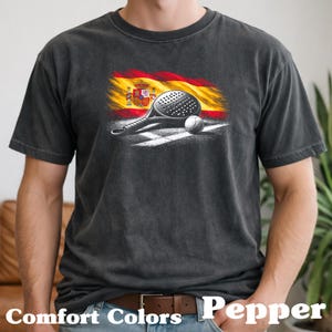 Könnte beinhalten: Dunkelgraues T-Shirt mit einer Grafik einer Padel-Schlägers, eines Balls und der spanischen Flagge. Das Design befindet sich vor einer weißen Spiellinie. Der Text "Comfort Colors Pepper" steht unten.