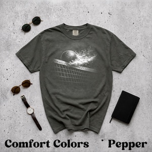 Könnte beinhalten: Dunkelgraues Comfort Colors T-Shirt mit einem Volleyball-Grafik. Das Design zeigt einen Volleyball, der über ein Netz geht, mit einem rauchigen Effekt. Sonnenbrillen, eine Uhr und ein Notizbuch sind ebenfalls im Bild.
