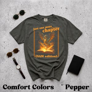 Könnte beinhalten: Dunkelgraues T-Shirt mit dem Text "just one more chapter (3AM edition)" in Orange. Die Grafik zeigt einen Drachen über einem offenen Buch mit Flammen. Sonnenbrillen, eine Uhr und ein kleines schwarzes Notizbuch sind ebenfalls zu sehen.
