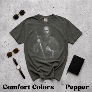 Könnte beinhalten: Pfefferfarbenes T-Shirt mit einer detaillierten Schwarz-Weiß-Illustration eines bärtigen Mannes, der ein Schwert und ein Buch hält. Der Text "Comfort Colors" und "Pepper" stehen unten. Sonnenbrillen und eine Uhr sind ebenfalls sichtbar.