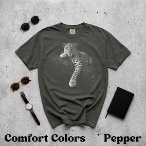 Könnte beinhalten: Dunkelgraues T-Shirt mit einer schwarz-weißen Illustration eines Leoparden auf einem Ast. Der Text "Comfort Colors Pepper" ist unten aufgedruckt. Sonnenbrillen, eine Uhr und ein kleines schwarzes Notizbuch sind ebenfalls zu sehen.