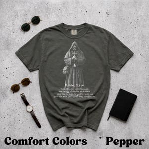 Könnte beinhalten: Pfefferfarbenes T-Shirt mit einer detaillierten Graustufenillustration einer Figur in Robe, die ein Kreuz hält. Der Text "Psalm 23:4" und ein Vers sind unter dem Bild aufgedruckt. Sonnenbrille, eine Uhr und ein kleines schwarzes Buch sind ebenfalls zu sehen.