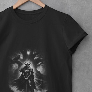 Könnte beinhalten: Schwarzes T-Shirt mit einer Graustufen-Illustration einer Kriegerin mit Hörnerhelm und Axt, vor einem dunklen Wald mit einem Raben. Das Design ist mittig auf dem T-Shirt platziert und erzeugt einen starken visuellen Kontrast.