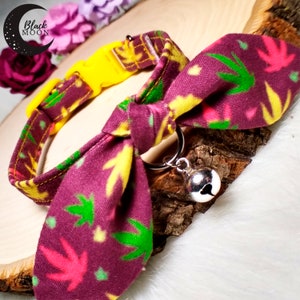 May include: A pet collar featuring a dark purple fabric with a repeating pattern of bright green, yellow, and pink leaf shapes. The collar includes a yellow plastic buckle, a tied fabric bow, a silver metal D-ring, and a small silver bell charm.