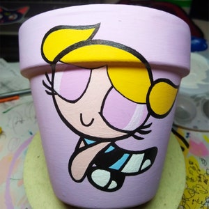 May include: A purple ceramic flower pot with a hand-painted cartoon image of a blonde girl with pink eyes and a blue and white striped outfit. The girl is smiling and has a yellow bow in her hair.