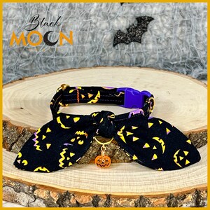 May include: A black fabric pet collar featuring a repeating yellow and purple jack-o'-lantern pattern, a purple plastic side-release buckle, a gold metal D-ring, and a small orange translucent pumpkin charm hanging from the center bow.