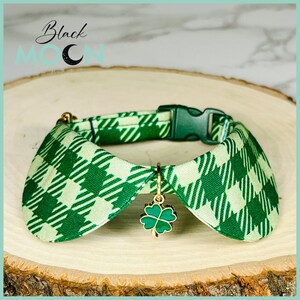 May include: A green and light green plaid fabric pet collar featuring a dark green plastic buckle and a gold-toned metal four-leaf clover charm. The collar has a rounded, folded collar-style design and is displayed on a circular wood slice.