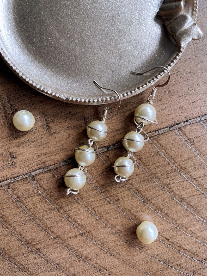 May include: A pair of pearl earrings with a silver wire design. The earrings are hanging from a silver tray.