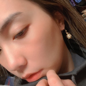 May include: A close-up of a person's face with a gold earring featuring a white and gold sphere pendant.