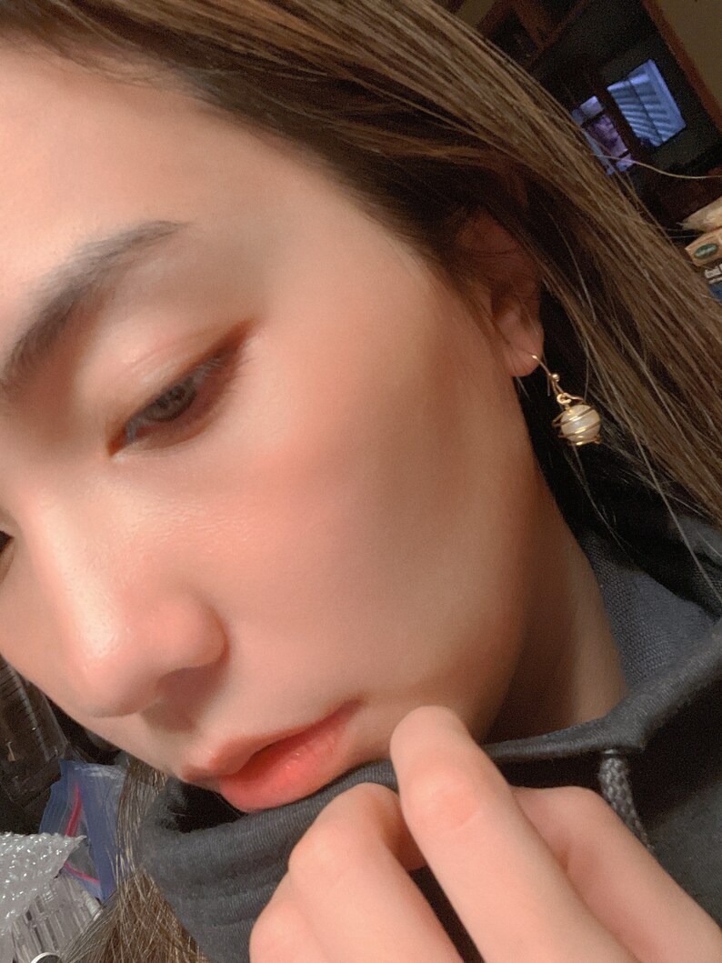 May include: A close-up of a person's face with a gold earring featuring a white and gold sphere pendant.
