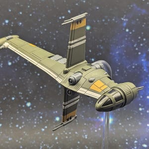 May include: A detailed model of a starfighter in shades of green, gray, and yellow. The spacecraft has a distinctive wing design and a cockpit with a clear canopy. The background features a starry space scene.