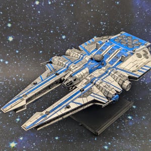 Puede incluir: Maqueta detallada de una nave espacial azul y plateada. La nave espacial tiene múltiples alas y motores, sobre un fondo estrellado. El modelo está montado sobre una base negra.