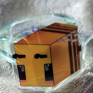 May include: A clear, glass-like ring with a cartoon bee design. The bee is yellow and brown with black accents, and is set within a clear, rounded setting. The ring is on a textured, white surface.