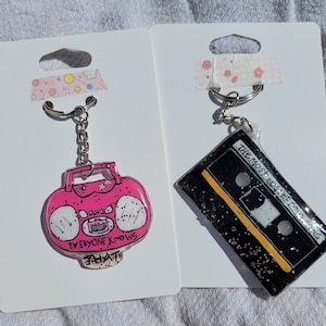 May include: Two keychains on white card stock. The keychain on the left is pink and features a cartoon boombox with the text "Everyone Knows That". The keychain on the right is black and features a cassette tape with the text "The Most Insincere Love Song".