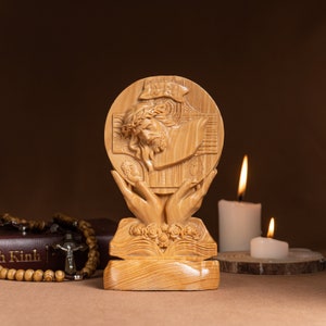 May include: A wooden sculpture of Jesus Christ on the cross, with a crown of thorns, surrounded by two hands holding a chalice and a host. The sculpture is on a wooden base with a book and flowers carved into it. The sculpture is in front of a brown background.