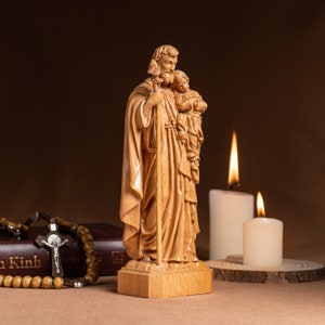 May include: A wooden statue of Saint Christopher carrying a child on his shoulder. The statue is standing on a wooden base and is surrounded by candles.