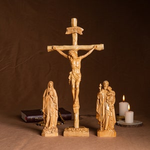 May include: A wooden crucifix with a carved figure of Jesus Christ, flanked by statues of the Virgin Mary and Saint Joseph. The statues are all made of light-colored wood and have a simple, traditional style. The crucifix is set on a wooden base and has a small inscription that reads "INRI".