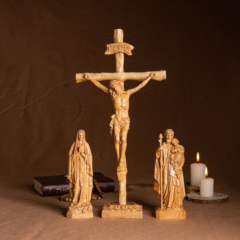 May include: A wooden crucifix with a carved figure of Jesus Christ, flanked by statues of the Virgin Mary and Saint Joseph. The statues are all made of light-colored wood and have a simple, traditional style. The crucifix is set on a wooden base and has a small inscription that reads "INRI".