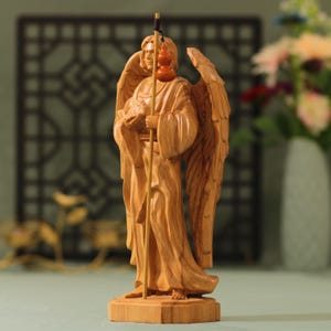 May include: A wooden sculpture of an angel with wings outstretched, holding a staff with gourds hanging from it. The angel is carved in a light brown wood and has a serene expression.