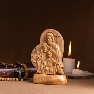 Op de afbeelding: Een houten beeldhouwwerk van de Heilige Familie, met Jezus, Maria en Jozef, in een hartvormig frame. Het beeldhouwwerk staat op een houten voet en heeft de woorden "God Bless Our Home" erop gegraveerd.