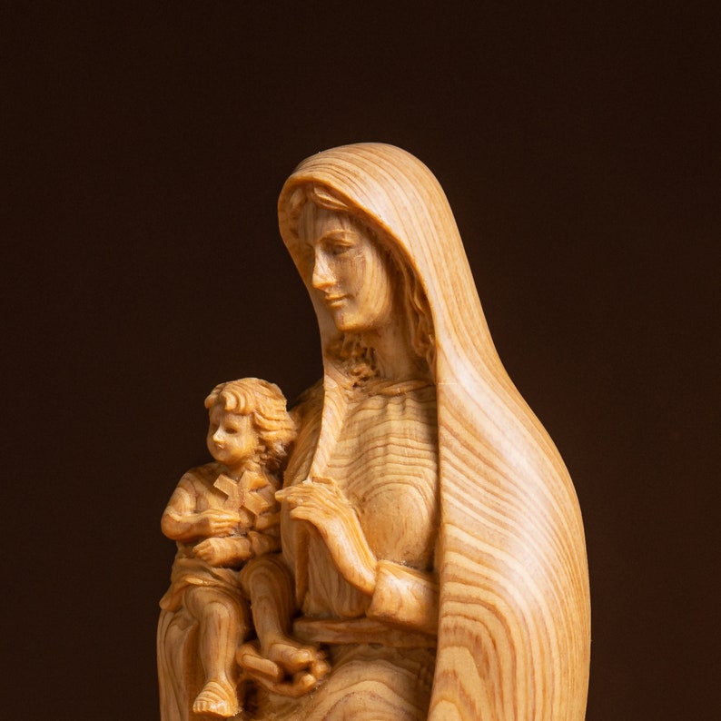 May include: A wooden sculpture of the Virgin Mary holding a child. The sculpture is carved from light brown wood with visible grain patterns. The Virgin Mary is wearing a long robe and a veil.