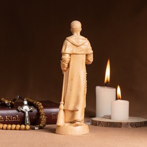 May include: A wooden figurine of a person in a robe, holding a broom and rosary beads. The figurine is standing on a wooden base. Two lit candles are in the background.