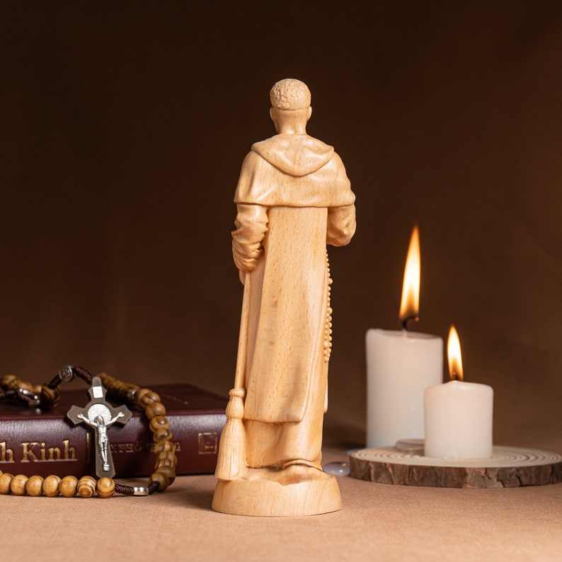 May include: A wooden figurine of a person in a robe, holding a broom and rosary beads. The figurine is standing on a wooden base. Two lit candles are in the background.