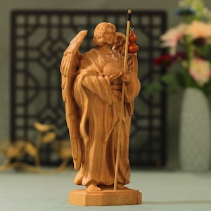 May include: A wooden sculpture of an angel with wings, holding a staff and a gourd. The angel is carved in a standing position with a flowing robe. The sculpture is made of light brown wood.