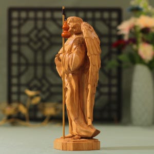 May include: A wooden sculpture of an angel with wings, holding a staff with gourds. The angel is carved in a light brown color and stands on a wooden base.