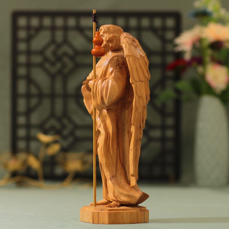 May include: A wooden sculpture of an angel with wings, holding a staff with gourds. The angel is carved in a light brown color and stands on a wooden base.