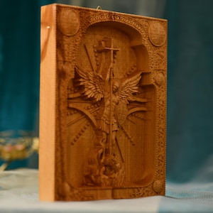 May include: A wooden carving of an angel with wings spread wide, holding a cross and standing on a serpent. The carving is surrounded by intricate details and a decorative frame.