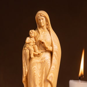 May include: A wooden statue of the Virgin Mary holding a baby Jesus. The statue is carved from light brown wood with a detailed grain pattern. The Virgin Mary is wearing a long robe and has a gentle expression on her face. The baby Jesus is sitting on her lap and is also carved from wood.