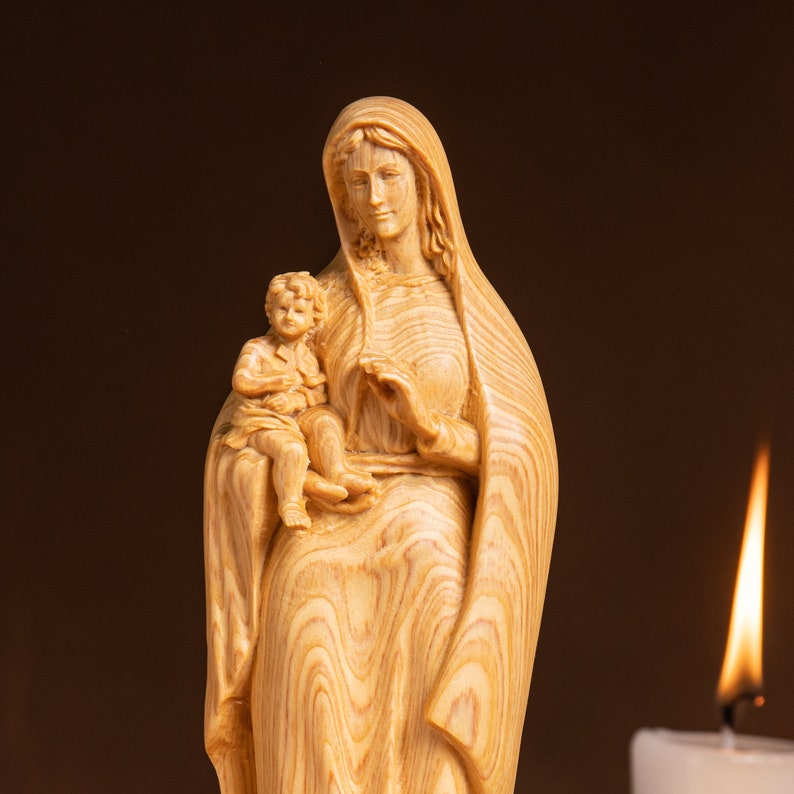 May include: A wooden statue of the Virgin Mary holding a baby Jesus. The statue is carved from light brown wood with a detailed grain pattern. The Virgin Mary is wearing a long robe and has a gentle expression on her face. The baby Jesus is sitting on her lap and is also carved from wood.