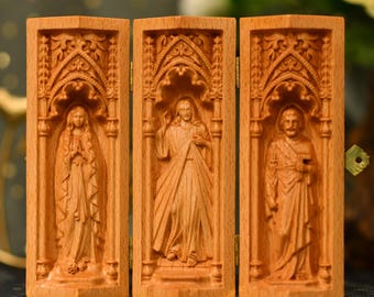 Könnte beinhalten: Ein hölzernes Triptychon mit geschnitzten religiösen Figuren. Die Scharnierplatten zeigen detaillierte Reliefschnitzereien von drei Figuren in gewölbten Rahmen. Das Holz hat einen warmen, natürlichen Farbton, und die Schnitzereien weisen komplizierte Details auf.