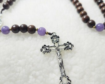 May include: A rosary with dark brown wooden beads and lavender-coloured beads. A silver crucifix with the figure of Jesus Christ is at the centre. The rosary is displayed on a white, textured surface.