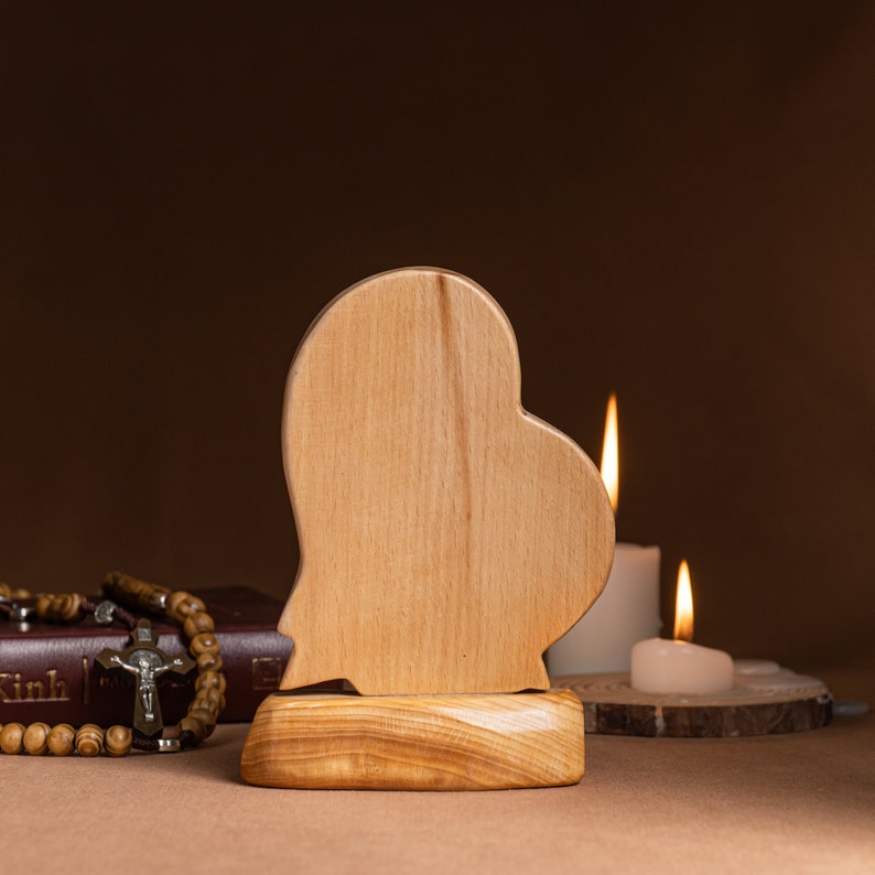 Op de afbeelding: Een houten hartvormige plaquette met een gladde, lichtbruine afwerking. De plaquette is gemonteerd op een houten basis met een natuurlijke houtnerf. Twee kaarsen zijn zichtbaar op de achtergrond, &eacute;&eacute;n brandend en &eacute;&eacute;n niet brandend.
