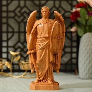 May include: A wooden sculpture of an angel with wings, holding a sword and a shield. The angel is carved in a standing position with a serious expression on its face.